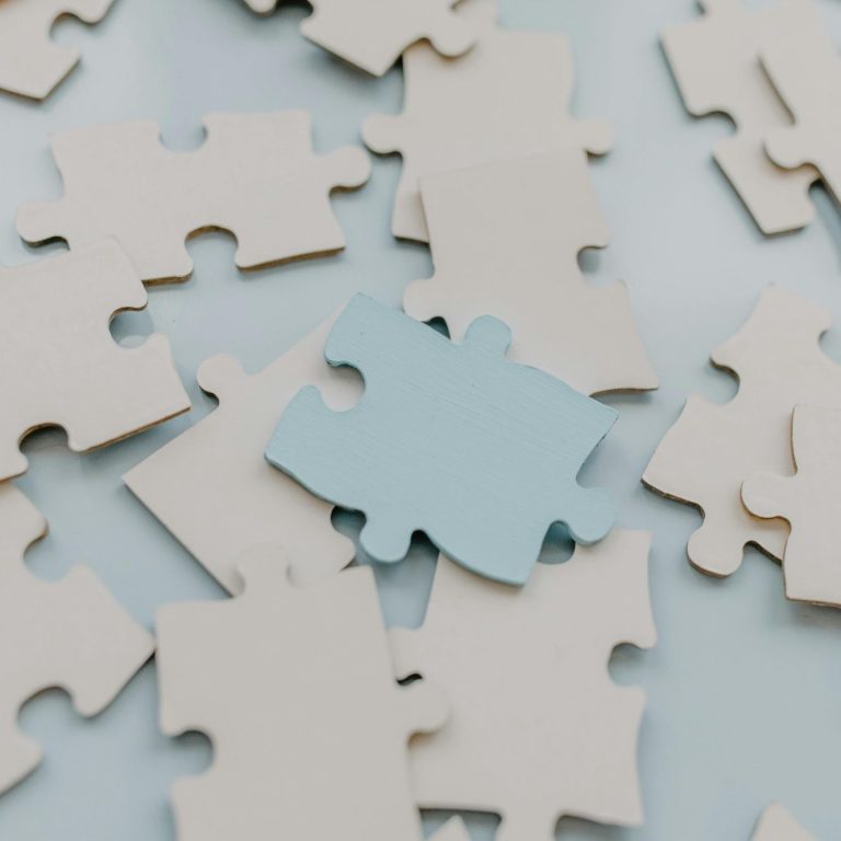 Kinder- und Jugendlichenpsychotherapie Mehrere helle Puzzleteile liegen ungeordnet auf einem Tisch, ein hellblaues Teil sticht hervor.